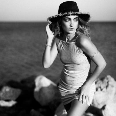 Black White Aufnahme einer Frau am Strand in einem knappen hellen Kleid und einem hut am Kopf.