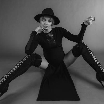 Black White Aufnahme einer blonden Frau in schwarz gekleidet mit Overknees. Sitz am Boden und schaut herausfordernd in die Kamera.