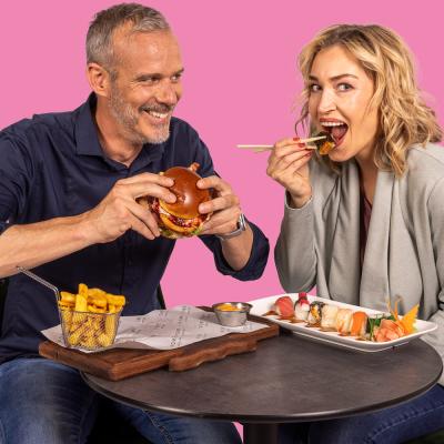 Mann sitzt mit Frau an Tisch mit rosa Hintergrund. Mann beisst in Burger und Frau hat Stäbchen und isst Sushi