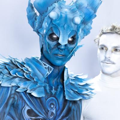 Bodypaintingkunst in blau. Sieht aus wie eine Mischung aus Mensch und Insekt. Daneben ein Mann ganz in weiß angemalen