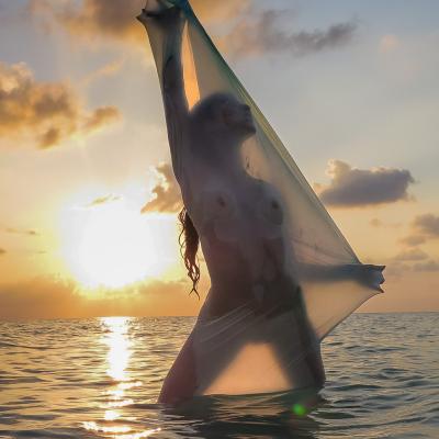 Frau steht im Meer. Im Hintergrund Sonne die untergeht. Frau Hält einen transparenten Stoff vor ihren Körper.