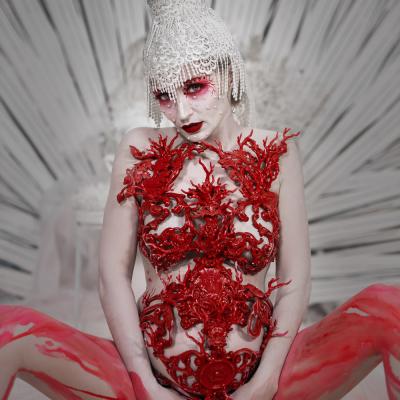 Ein beeindruckendes künstlerisches Porträt einer Person mit heller Haut, die eine aufwendige weiße Kopfbedeckung und dramatisches rotes Make-up trägt