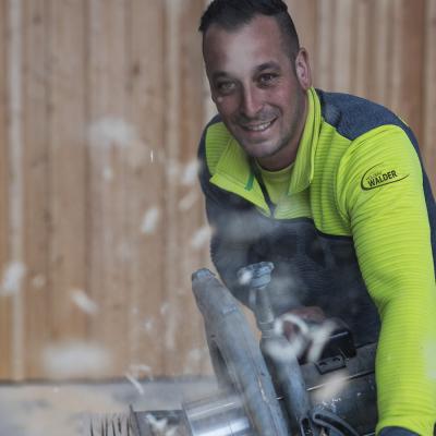 Mann in gelb-grauer Jacke an Holzschleifmaschine lächelt in die Kamera