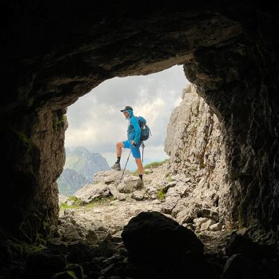 Mann in blauen Wanderkleidung und mit Rucksack und Cappi steht auf Felsen und wird von der Kamera durch einen Höhlenausgang aufgenommen
