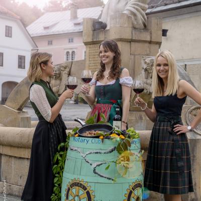 drei junge Frauen im Dirndl stehen vor Brunnen und hinter einem türkisen Weinfass und stoßen mit Rotwein an