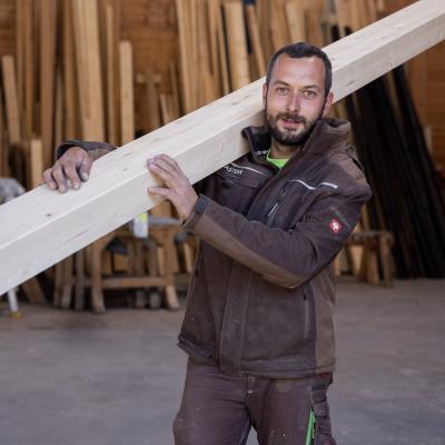 Mann mit dunklem Bart und Arbeitsmontur trägt Holzträger und schaut in die Kamera