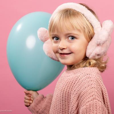 Blondes Mädchen in Rosa gekleidet, haltet einen türkisen Luftballon in der Hand und lächelt in die Kamera 