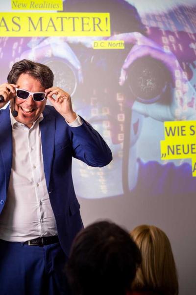 Ein Mann in blauem Anzug steht vor einem Publikum und setzt sich lächelnd eine weiße Sonnenbrille auf. Hinter ihm ist eine bunte Präsentationsfolie projiziert mit den Texten „IDEAS MATTER!“ und „Wie sieht das ‚neue Denken‘ aus?“. 