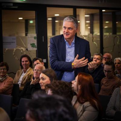 Ein Mann im blauen Sakko steht inmitten eines Publikums in einem Konferenzraum auf und spricht engagiert, während sich die übrigen Teilnehmenden aufmerksam umdrehen 