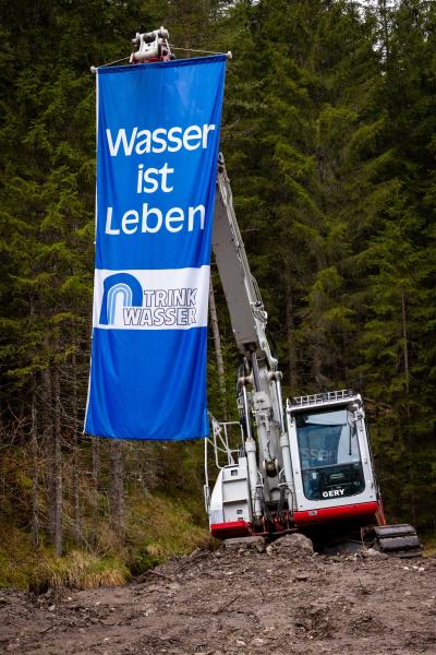 Ein Bagger auf einer matschigen Baustelle inmitten eines Waldes hält ein großes blaues Banner mit der Aufschrift „Wasser ist Leben“