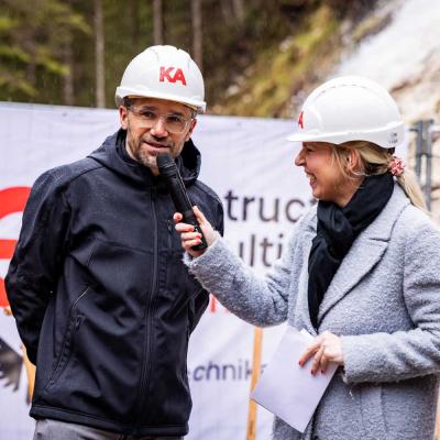 Ein Mann und eine Frau mit weißen Bauhelmen und wetterfester Kleidung bei einer Baustelleneröffnung