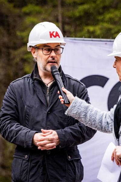 Ein Mann mit Brille und weißem Bauhelm wird bei einer Veranstaltung im Freien interviewt. Er trägt eine dunkle Jacke und spricht in ein Mikrofon, das ihm von einer Frau mit ebenfalls weißem Bauhelm und grauem Mantel entgegengehalten wird.