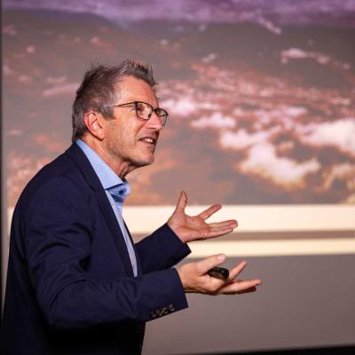 Ein Mann mittleren Alters mit Brille und Anzug spricht gestikulierend während einer Präsentation vor einer großen Leinwand