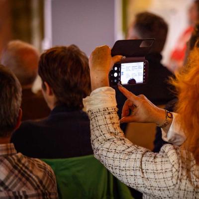Eine Frau mit auffällig roten Haaren fotografiert mit ihrem Smartphone eine Präsentation oder Folie während einer Veranstaltung. 