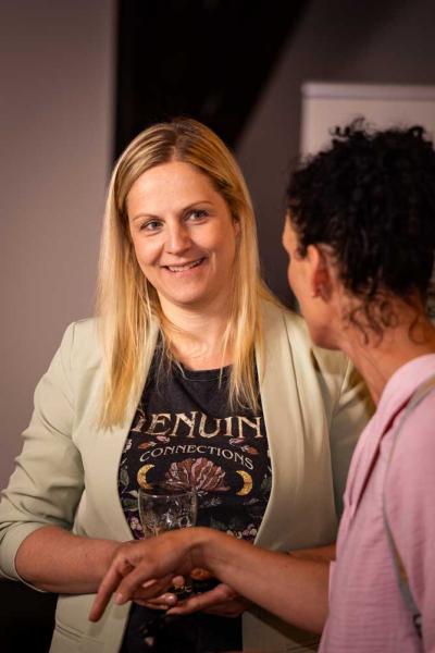 Zwei Frauen unterhalten sich freundlich in einem Innenraum. Die blonde Frau im Vordergrund lächelt, hält ein leeres Trinkglas und trägt ein T-Shirt mit dem Aufdruck „Genuine Connections“. Die andere Frau trägt einen Rucksack und ist von hinten zu sehen.