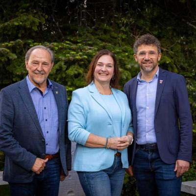 Ein Gruppenfoto von drei Personen – zwei Männer und eine Frau – die nebeneinander im Freien vor grünen Tannen stehen. 